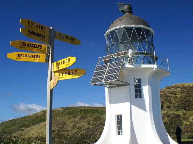 Nahaufnahme des Leuchtturms von Cape Reinga und eines gelben Wegweisers mit Entfernungen zu verschiedenen Städten.