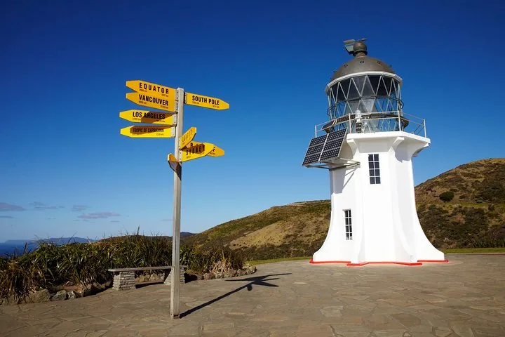 Der Leuchtturm von Cape Reinga und der ikonische gelbe Entfernungswegweiser an einem sonnigen Tag.