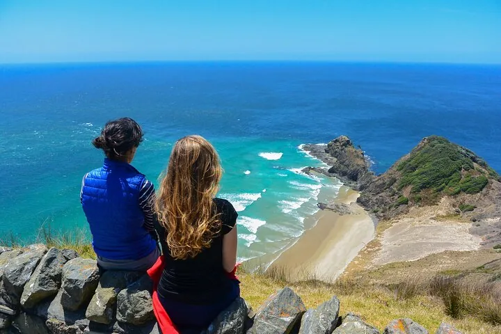Zwei Personen sitzen auf einer Steinmauer und blicken in Cape Reinga aufs Meer hinaus.